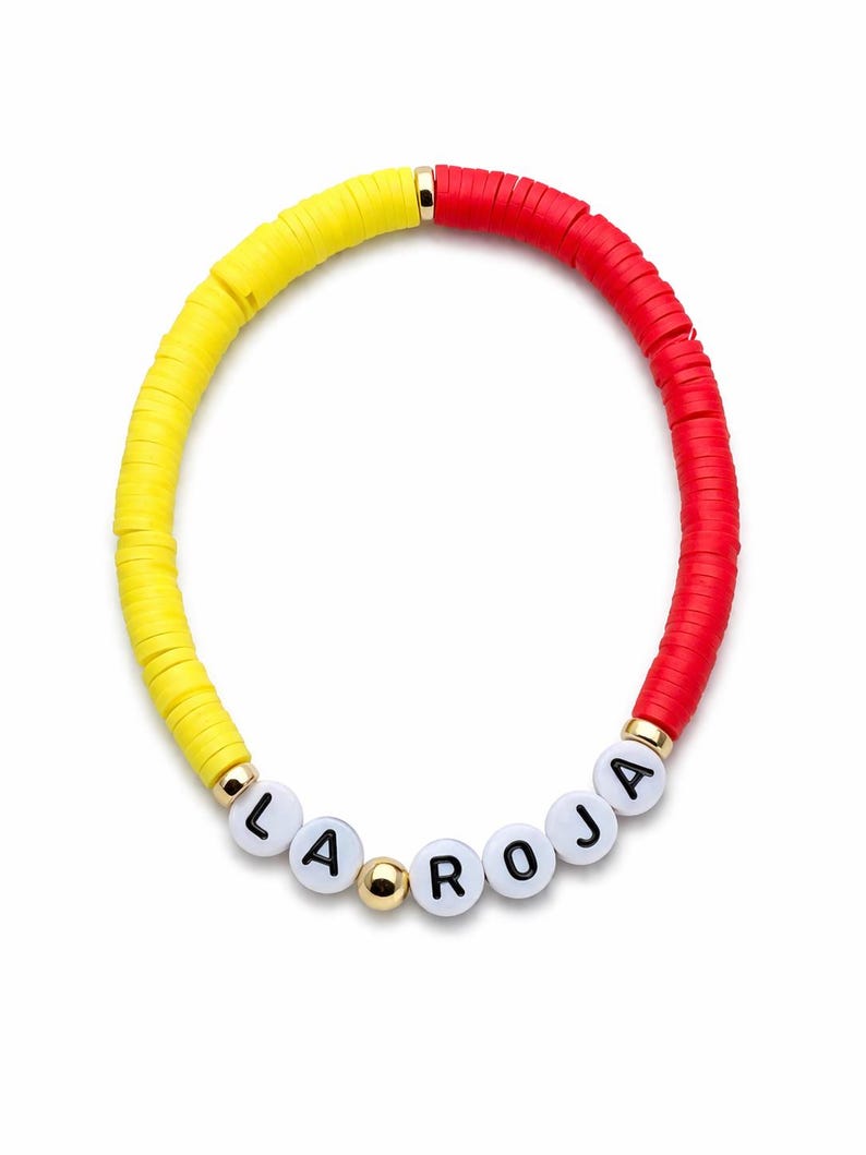 May include: A colorful bracelet featuring yellow and red flat beads. The bracelet includes white letter beads that spell out "LA ROJA" and gold-colored accents. The bracelet is a vibrant accessory.
