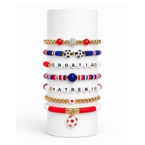 May include: A collection of beaded bracelets in red, white, blue, and gold. Some bracelets feature the word "CROATIA" and "VATRENI" spelled out with letter beads. Other bracelets have soccer ball charms and beads.