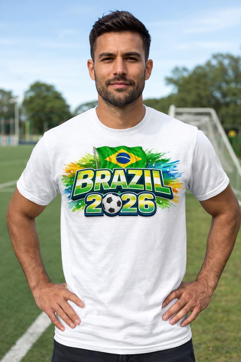 May include: White t-shirt featuring the text "BRAZIL 2026" in green and yellow, a football, and the Brazilian flag. The shirt has a colourful design and is worn by a person standing on a green field.