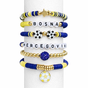 May include: A collection of beaded bracelets in blue, yellow, and gold. Some bracelets spell out "BOSNA" and "HERCEGOVINA" with white letter beads. Other bracelets feature soccer ball charms and star-shaped accents.