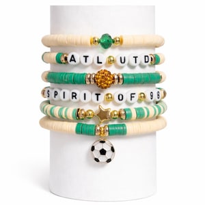 Puede incluir: Conjunto de pulseras de cuentas en crema, verde y dorado. Algunas pulseras deletrean "ATLUTD" y "SPIRIT OF 98" con cuentas de letras blancas. Un dije de balón de fútbol cuelga de una pulsera.