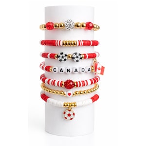 May include: A stack of seven beaded bracelets in red, white, and gold. Features include a crystal-encrusted bead, soccer ball beads, white letter beads spelling CANADA, a Canadian flag charm, a red glitter bead, a heart bead, and a soccer ball charm.