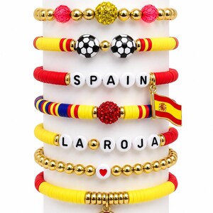 May include: A collection of colorful beaded bracelets with a Spanish theme. The bracelets feature gold beads, soccer balls, the word "SPAIN", the phrase "LA ROJA", and a Spanish flag charm. The bracelets are in red, yellow, and blue.