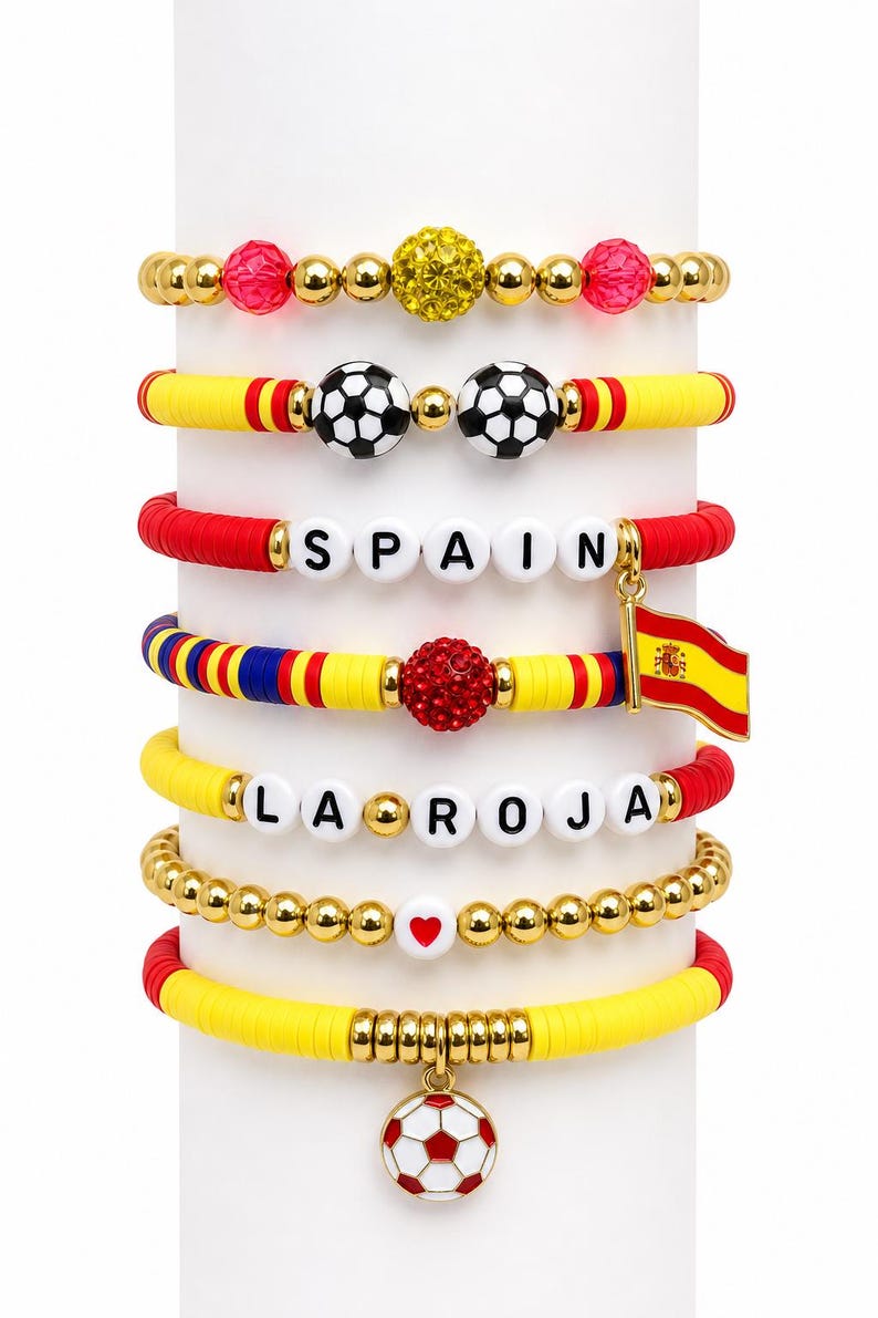 May include: A collection of colorful beaded bracelets with a Spanish theme. The bracelets feature gold beads, soccer balls, the word "SPAIN", the phrase "LA ROJA", and a Spanish flag charm. The bracelets are in red, yellow, and blue.