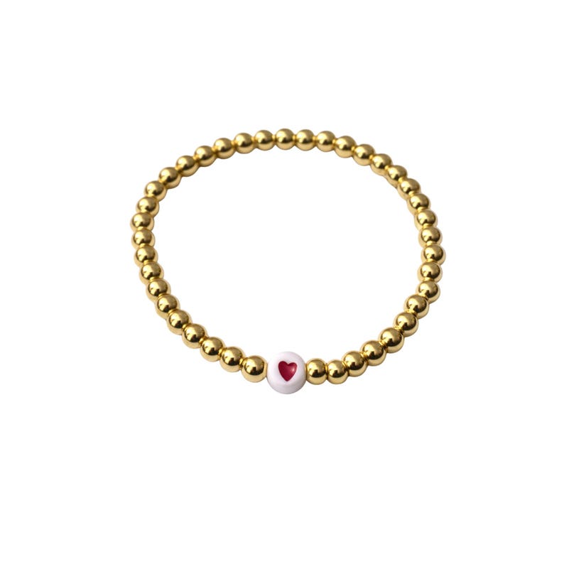 May include: Gold beaded bracelet with a white heart-shaped bead featuring a red heart design. The bracelet is made of small, round, gold-colored beads. The heart bead is centered on the bracelet.