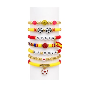 Op de afbeelding: Een stapel van zeven kralenarmbanden met goudkleurige kralen, rode en gele kleischijfjes en voetbalbedels. Eén armband spelt SPAIN met een Spaanse vlagbedel, een andere spelt LA ROJA en één armband heeft een voetbalhanger als detail.