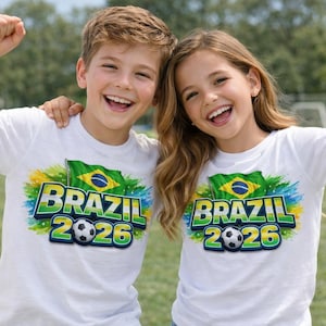 May include: Two children wearing white t-shirts with a colourful Brazil 2026 graphic. The design includes the Brazilian flag, the word "BRAZIL", the year "2026", and a football. The children are smiling and raising their arms.