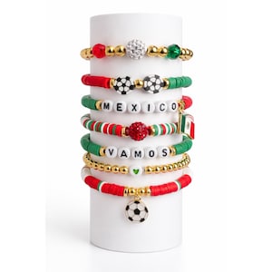 Puede incluir: Una colección de pulseras de cuentas rojas, verdes, blancas y doradas, con dijes de balón de fútbol y bandera. Algunas pulseras tienen las palabras "MEXICO" y "VAMOS" deletreadas en cuentas blancas. Las pulseras se exhiben en un cilindro blanco.
