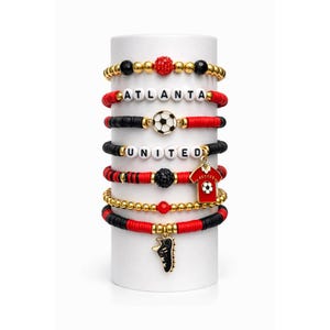 以下が含まれることがあります： サッカーをテーマにしたチャームが付いた重ね付けブレスレット。ブレスレットは赤、黒、金色のビーズで、サッカーボール、ジャージ、クリートなどのチャームが付いています。「ATLANTA」と「UNITED」と書かれたブレスレットもあります。