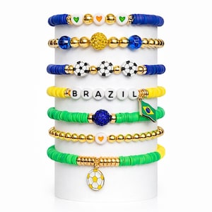 May include: Stack of colorful beaded bracelets with soccer ball and Brazil flag charms. The bracelets feature blue, yellow, green, and gold beads, with the word "BRAZIL" spelled out in white letter beads.