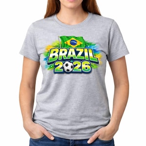 May include: A grey t-shirt featuring the text "BRAZIL 2026" in green and blue, a football, and the Brazilian flag. The design has a colourful splash background. The shirt is a crew neck style.
