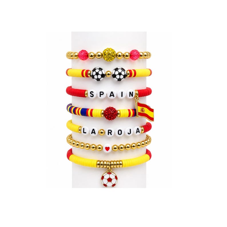 May include: A collection of colorful beaded bracelets. The bracelets feature gold, red, yellow, and blue beads, with soccer ball charms, the word "SPAIN", and a Spanish flag charm. Some bracelets have heart-shaped beads.