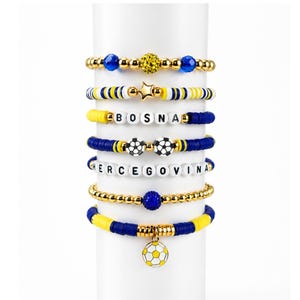 May include: A selection of beaded bracelets in gold, blue, and yellow. Some bracelets feature soccer ball charms, star charms, and the words "BOSNA" and "HERCEGOVINA" spelled out in white beads. The bracelets are displayed on a white cylinder.