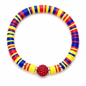 May include: A colorful bracelet featuring alternating red, yellow, and blue disc-shaped beads. A central red, crystal-studded ball is accented with gold-colored metal rings. The bracelet is designed to be worn around the wrist.