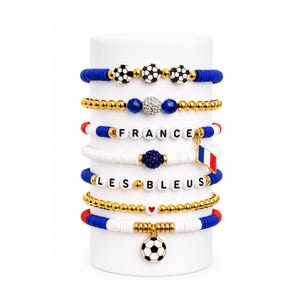 May include: A set of seven stacked bracelets featuring blue, white, and gold beads. The bracelets include soccer ball charms, a French flag charm, and white letter beads spelling FRANCE and LES BLEUS, accented with gold spheres and a small red heart.