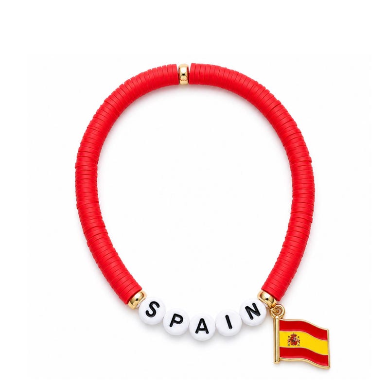 May include: A red beaded bracelet with the word "SPAIN" spelled out in white letter beads. A gold-colored charm of the Spanish flag hangs from the bracelet. The bracelet is on a white background.