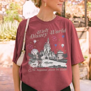 A woman wearing a red t-shirt with a Disney World graphic and text, standing in front of a brick wall with greenery.