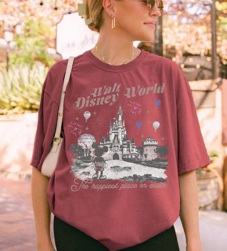 A woman wearing a red t-shirt with a Disney World graphic and text, standing in front of a brick wall with greenery.