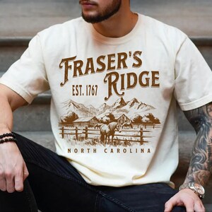 A man wearing a white t-shirt with a graphic design of a mountain landscape and the text "Fraser's Ridge" printed on it.