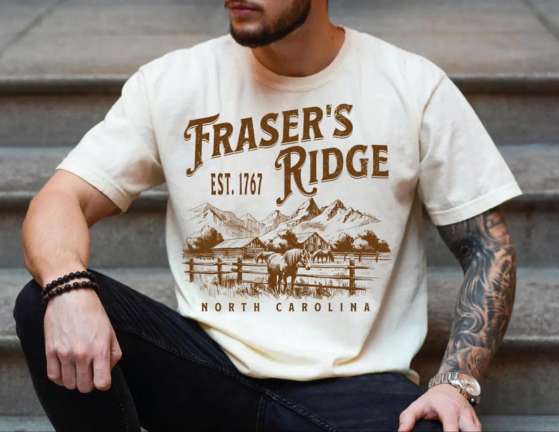 A man wearing a white t-shirt with a graphic design of a mountain landscape and the text "Fraser's Ridge" printed on it.