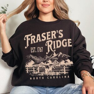 A woman wearing a black sweatshirt with a graphic design of a mountain range and the text "Fraser's Ridge" printed on it.