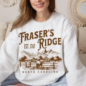 A woman wearing a white sweatshirt with a graphic design depicting a mountain landscape and the text "Fraser's Ridge" printed on it.