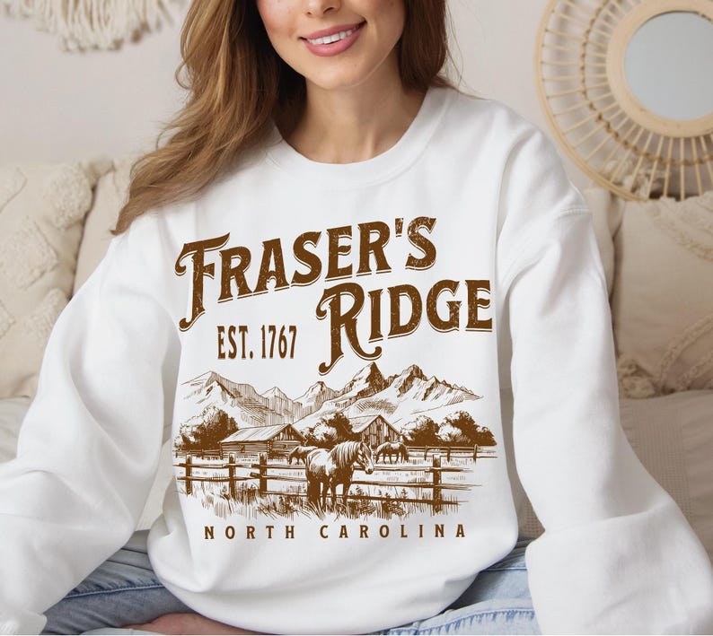 A woman wearing a white sweatshirt with a graphic design depicting a mountain landscape and the text "Fraser's Ridge" printed on it.