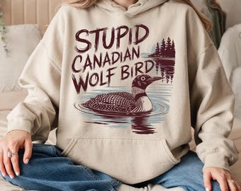 May include: Beige hoodie with the text "STUPID CANADIAN WOLF BIRD" in maroon. The graphic features a loon bird in water with a forest and lake scene. The hoodie has a drawstring hood and a front pocket.