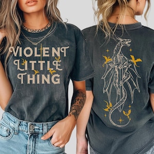 May include: Dark gray t-shirt with the text "VIOLENT LITTLE THING" in a vintage font. The back features a dragon design with a sword, wings, and yellow accents. A casual, graphic tee.