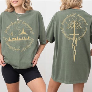 a woman wearing a green t-shirt with a graphic design featuring a castle, a tree, and a sword. The design is symmetrical, with the castle and tree on the left side and the sword on the right side. The woman is posing with her hands on her hips, showcasing the t-shirt design.