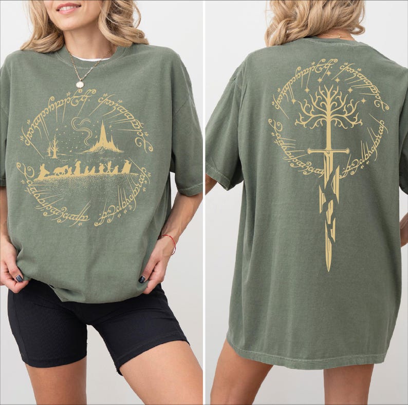 a woman wearing a green t-shirt with a graphic design featuring a castle, a tree, and a sword. The design is symmetrical, with the castle and tree on the left side and the sword on the right side. The woman is posing with her hands on her hips, showcasing the t-shirt design.