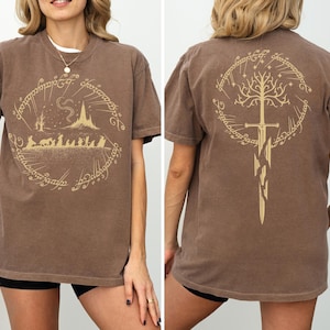 a woman wearing a brown t-shirt with a graphic design featuring a castle, a tree, and a sword. The design is symmetrical, with the castle and tree on the left and the sword on the right.
