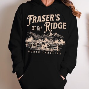 A black hoodie with a graphic design depicting a mountain range and a horse, along with text that reads "Fraser's Ridge Est. 1767 North Carolina".