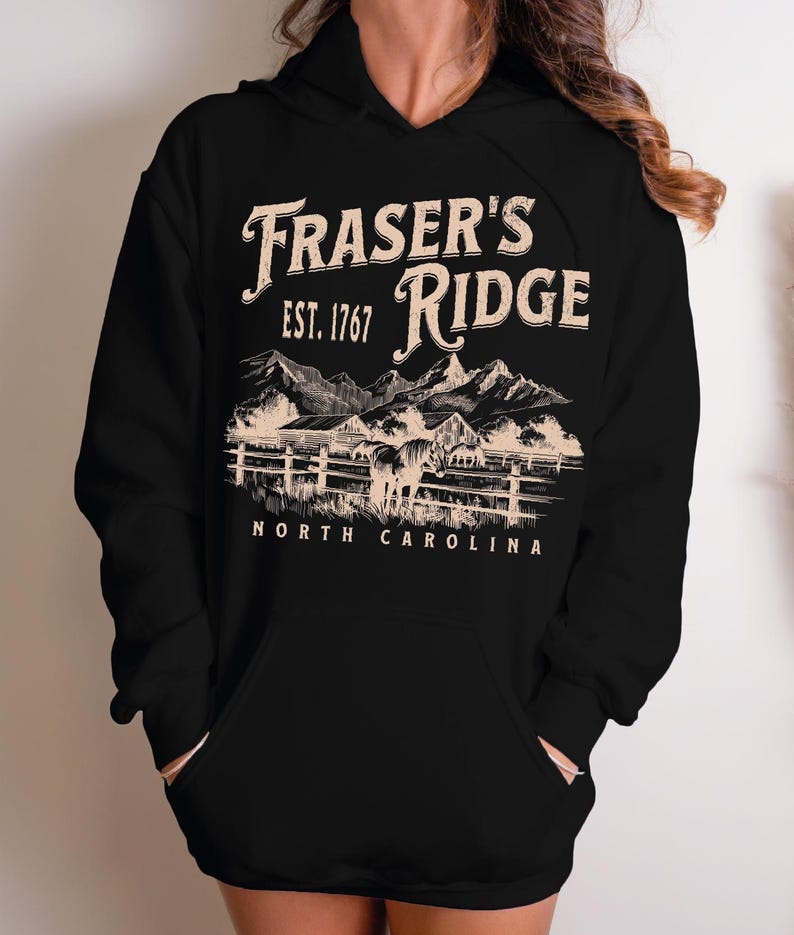 A black hoodie with a graphic design depicting a mountain range and a horse, along with text that reads "Fraser's Ridge Est. 1767 North Carolina".