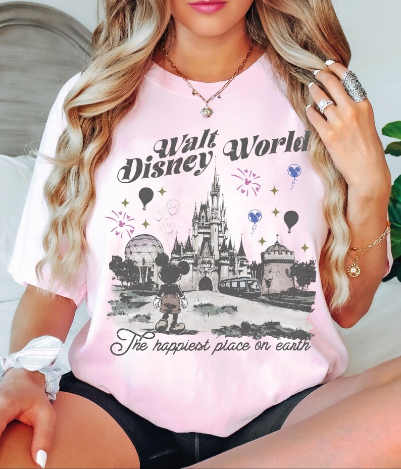 A woman wearing a pink t-shirt with a Disney World graphic and text, posing in front of a bed.