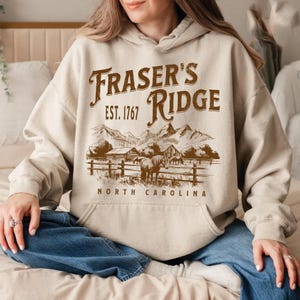 A woman wearing a beige hoodie with a graphic design of a mountain range and the text "Fraser's Ridge" printed on it.