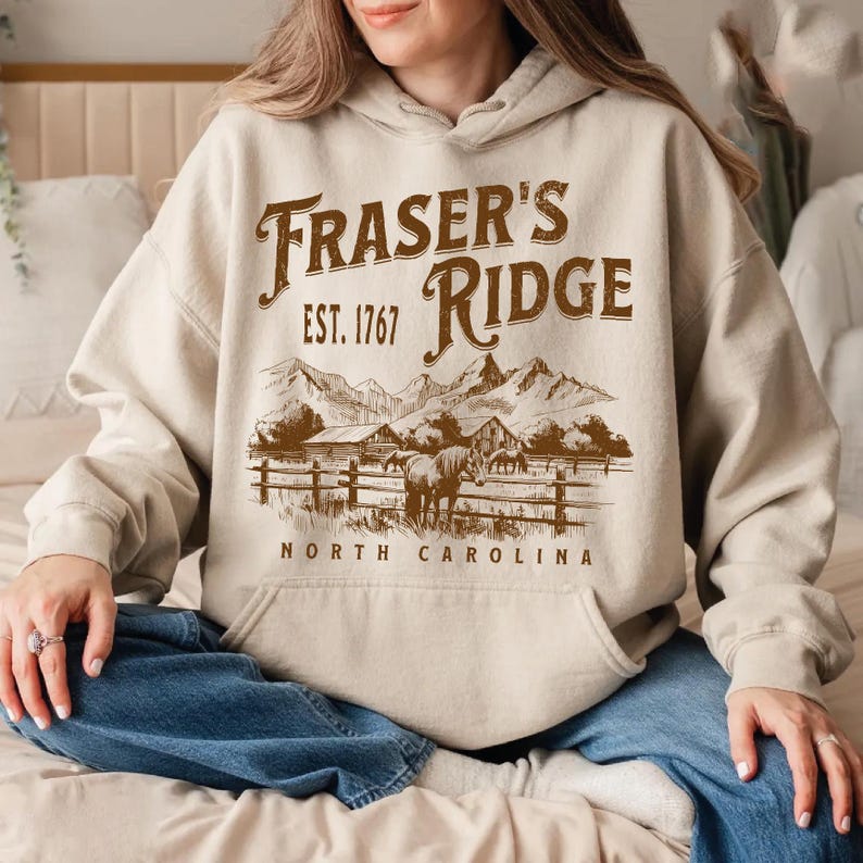 A woman wearing a beige hoodie with a graphic design of a mountain range and the text "Fraser's Ridge" printed on it.
