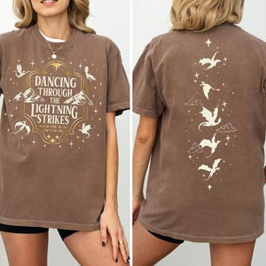 A woman wearing a brown t-shirt with a design featuring a dancing dragon and the text "Dancing through the lightning strikes".
