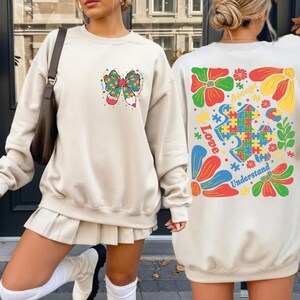 May include: Beige sweatshirt with a colorful bow design on the front and a graphic on the back. The back graphic includes puzzle pieces, flowers, and the words "Love, Accept, Understand."