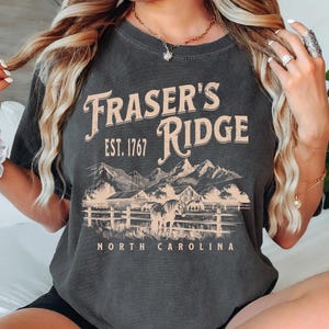 A woman wearing a gray t-shirt with the text "Fraser's Ridge" and "Est. 1767 North Carolina" printed on it.