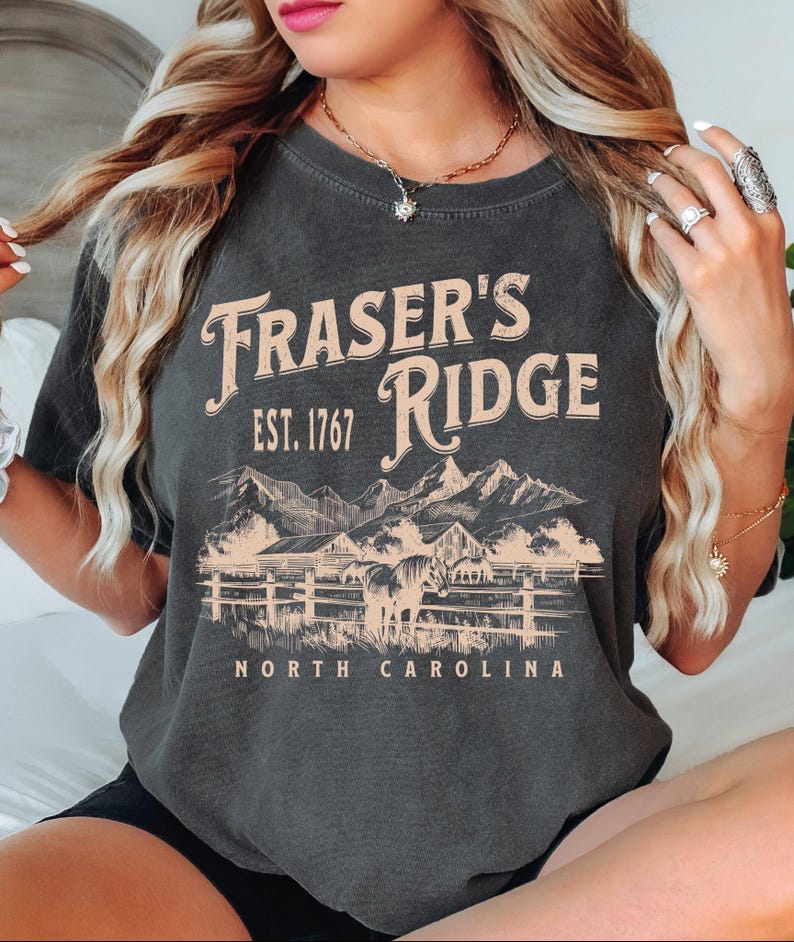 A woman wearing a gray t-shirt with the text "Fraser's Ridge" and "Est. 1767 North Carolina" printed on it.