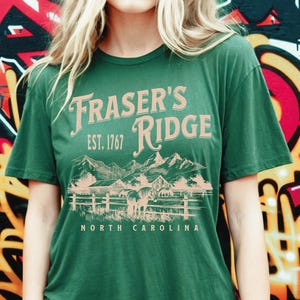 A woman wearing a green t-shirt with the text "Fraser's Ridge" and "Est. 1767" printed on it, standing in front of a colorful graffiti-covered wall.