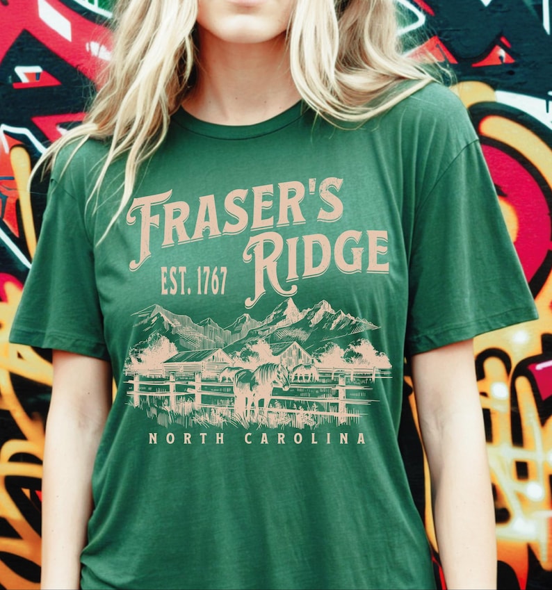 A woman wearing a green t-shirt with the text "Fraser's Ridge" and "Est. 1767" printed on it, standing in front of a colorful graffiti-covered wall.