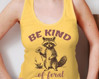May include: A yellow racerback tank top featuring a dark brown graphic of a raccoon holding a daisy. The text above the raccoon reads BE KIND and the text below reads ...of feral in a dark brown font. The design is centered on the front.
