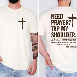May include: Cream-colored t-shirt with a brown cross on the front and back. The back of the shirt has the text "NEED PRAYER? TAP MY SHOULDER." and a Bible verse from Matthew 18:20.