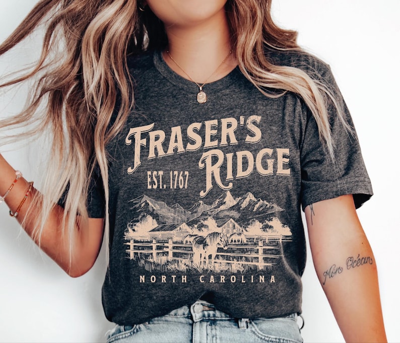 A woman wearing a gray t-shirt with the text "Fraser's Ridge" and "North Carolina" printed on it, along with a mountain landscape graphic.