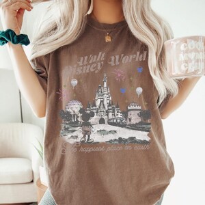 A woman wearing a brown t-shirt with a Disney World-themed graphic and text, holding a mug with a similar design.