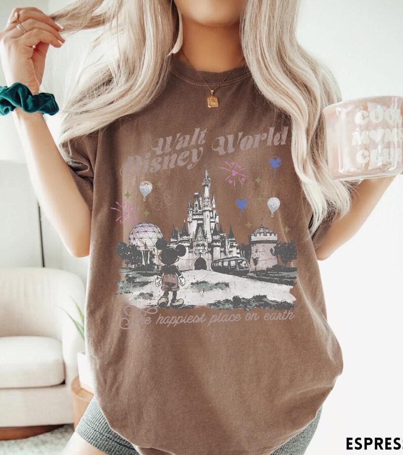 A woman wearing a brown t-shirt with a Disney World-themed graphic and text, holding a mug with a similar design.