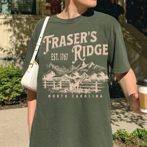 A woman wearing a green t-shirt with the text "Fraser's Ridge" and "Est. 1767" printed on it, standing outdoors in front of a building.
