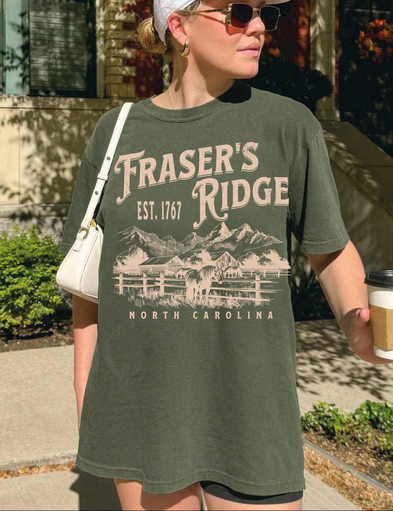 A woman wearing a green t-shirt with the text "Fraser's Ridge" and "Est. 1767" printed on it, standing outdoors in front of a building.